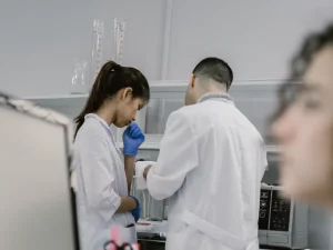 Dua peneliti berjas laboratorium bekerja di dekat instrumen, sementara seorang lainnya mencatat di depan komputer