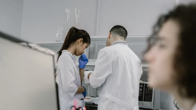 Dua peneliti berjas laboratorium bekerja di dekat instrumen, sementara seorang lainnya mencatat di depan komputer