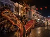 Ilustrasi seorang penampil mengenakan kostum karnaval bersayap lebar berhias bulu dan untaian lampu kecil dengan hiasan kepala menyerupai kepala naga, berarak di jalan kota berbangunan kolonial pada malam hari