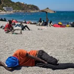 Seorang pria berkaus oranye tertidur telungkup di pantai berkerikil dengan puluhan orang lain di kejauhan