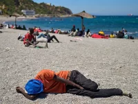 Seorang pria berkaus oranye tertidur telungkup di pantai berkerikil dengan puluhan orang lain di kejauhan