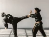 Ilustrasi dua atlet bela diri berseragam hitam bersabuk berlatih tendangan tinggi dan tangkisan di ruang latihan