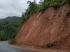 Lereng galian tanah residual kemerahan di tepi jalan perbukitan setelah hujan.