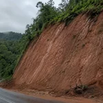 Lereng galian tanah residual kemerahan di tepi jalan perbukitan setelah hujan.