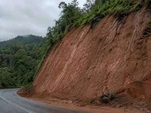 Lereng galian tanah residual kemerahan di tepi jalan perbukitan setelah hujan.