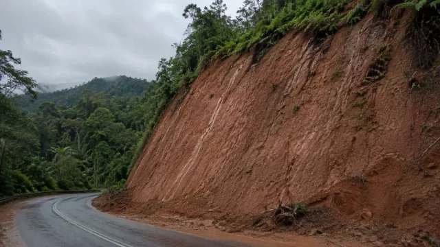 Lereng galian tanah residual kemerahan di tepi jalan perbukitan setelah hujan.
