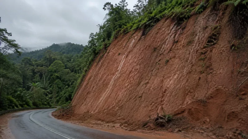Lereng galian tanah residual kemerahan di tepi jalan perbukitan setelah hujan.