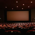 Deretan kursi merah menghadap layar bioskop yang menyala di ruang gelap