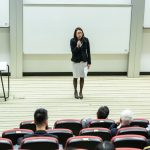 Ilustrasi seorang pembicara menyampaikan materi di hadapan peserta seminar pendidikan.