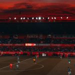 Ilustrasi laga sepak bola di stadion saat matahari terbenam.