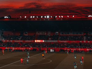 Ilustrasi laga sepak bola di stadion saat matahari terbenam.
