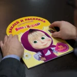 Seseorang memegang buku stiker serial animasi Masha and the Bear di atas meja kayu.
