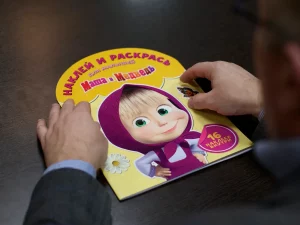 Seseorang memegang buku stiker serial animasi Masha and the Bear di atas meja kayu.