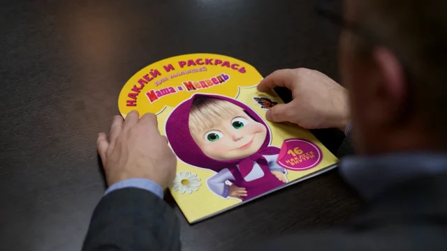 Seseorang memegang buku stiker serial animasi Masha and the Bear di atas meja kayu.