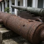 Ilustrasi meriam besi tua berkarat di atas landasan batu dengan lubang sundut terlihat jelas, berlatar meriam perunggu berpatina hijau di atas kereta kayu jati di beranda gedung museum bergaya kolonial