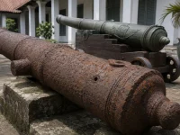 Ilustrasi meriam besi tua berkarat di atas landasan batu dengan lubang sundut terlihat jelas, berlatar meriam perunggu berpatina hijau di atas kereta kayu jati di beranda gedung museum bergaya kolonial