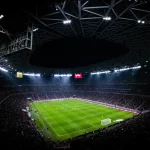 Ilustrasi pertandingan sepak bola malam hari yang disaksikan penonton di stadion.
