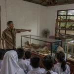 Ilustrasi ruang pamer museum komunitas di pedesaan Jawa Tengah dengan lemari kaca dan rak kayu berisi fosil rahang serta tulang, seorang pemandu berkemeja batik menunjuk vitrin, dan rombongan murid berseragam menyimak