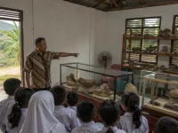 Ilustrasi ruang pamer museum komunitas di pedesaan Jawa Tengah dengan lemari kaca dan rak kayu berisi fosil rahang serta tulang, seorang pemandu berkemeja batik menunjuk vitrin, dan rombongan murid berseragam menyimak