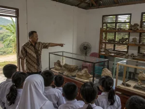 Ilustrasi ruang pamer museum komunitas di pedesaan Jawa Tengah dengan lemari kaca dan rak kayu berisi fosil rahang serta tulang, seorang pemandu berkemeja batik menunjuk vitrin, dan rombongan murid berseragam menyimak