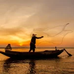 Seorang nelayan berdiri di perahu kayu sambil menebar jala di permukaan air saat matahari terbenam