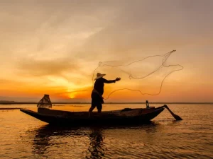 Seorang nelayan berdiri di perahu kayu sambil menebar jala di permukaan air saat matahari terbenam