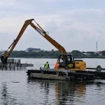 Ekskavator amfibi mengeruk dasar Waduk Pusong ditemani dua pekerja berrompi keselamatan