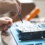 Sepasang tangan memperbaiki papan sirkuit elektronik di atas meja kerja dengan peralatan servis
