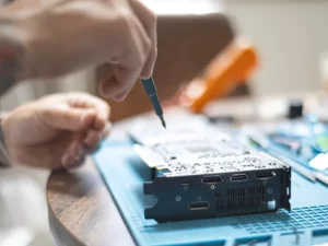 Sepasang tangan memperbaiki papan sirkuit elektronik di atas meja kerja dengan peralatan servis