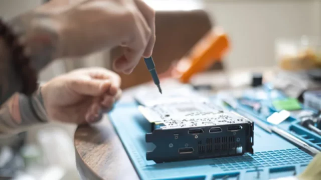 Sepasang tangan memperbaiki papan sirkuit elektronik di atas meja kerja dengan peralatan servis
