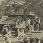 Foto hitam putih pasar Payakumbuh sekitar 1910 dengan los beratap rumbia, pedagang duduk beralas tikar, dan keranjang hasil bumi di tanah