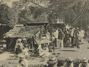 Foto hitam putih pasar Payakumbuh sekitar 1910 dengan los beratap rumbia, pedagang duduk beralas tikar, dan keranjang hasil bumi di tanah