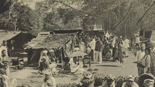Foto hitam putih pasar Payakumbuh sekitar 1910 dengan los beratap rumbia, pedagang duduk beralas tikar, dan keranjang hasil bumi di tanah