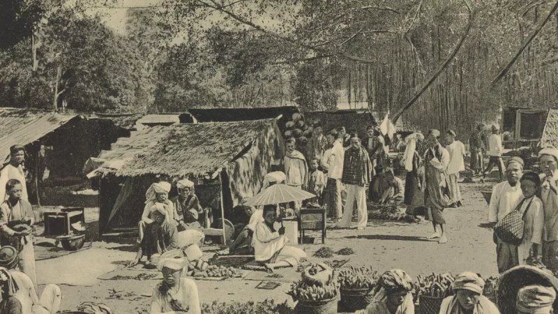 Foto hitam putih pasar Payakumbuh sekitar 1910 dengan los beratap rumbia, pedagang duduk beralas tikar, dan keranjang hasil bumi di tanah