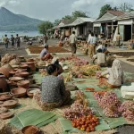 Ilustrasi pasar terbuka di tepi pantai berpasir gelap dengan gerabah tanah liat, ikatan bawang, daun pisang berisi hasil kebun, tikar penjemuran cengkeh, deretan ruko kayu beratap seng, dan perahu bercadik di latar gunung berapi