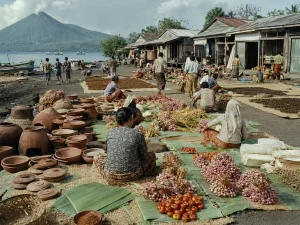 Ilustrasi pasar terbuka di tepi pantai berpasir gelap dengan gerabah tanah liat, ikatan bawang, daun pisang berisi hasil kebun, tikar penjemuran cengkeh, deretan ruko kayu beratap seng, dan perahu bercadik di latar gunung berapi