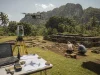 Ilustrasi drone melayang di atas teras batu situs candi yang sedang digali, dengan alat ukur total station bertripod, layar model permukaan tanah, dan dua pekerja lapangan bertopi lebar