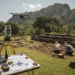 Ilustrasi drone melayang di atas teras batu situs candi yang sedang digali, dengan alat ukur total station bertripod, layar model permukaan tanah, dan dua pekerja lapangan bertopi lebar