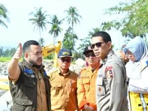Sejumlah petugas berseragam meninjau kawasan sungai berbatu yang terdampak banjir dengan alat berat di latar belakang
