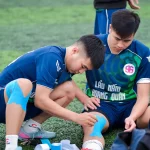 Ilustrasi dua pemain sepak bola muda memasang plester kinesiologi pada lutut dan pergelangan kaki di tepi lapangan