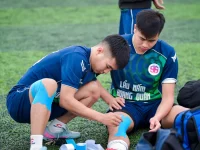 Ilustrasi dua pemain sepak bola muda memasang plester kinesiologi pada lutut dan pergelangan kaki di tepi lapangan