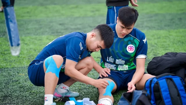 Ilustrasi dua pemain sepak bola muda memasang plester kinesiologi pada lutut dan pergelangan kaki di tepi lapangan