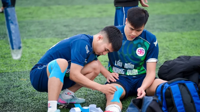 Ilustrasi dua pemain sepak bola muda memasang plester kinesiologi pada lutut dan pergelangan kaki di tepi lapangan