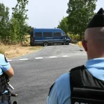 Dua petugas gendarmerie berompi antipeluru berjaga di persimpangan jalan dengan mobil van polisi di kejauhan