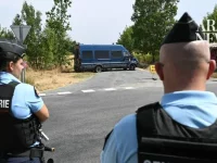 Dua petugas gendarmerie berompi antipeluru berjaga di persimpangan jalan dengan mobil van polisi di kejauhan