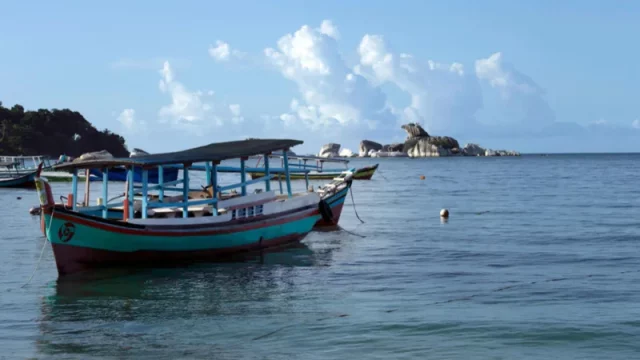 Perahu nelayan kayu tradisional berlabuh di laut tenang dengan gugusan batu granit dan pulau kecil di kejauhan