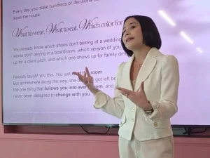 Prilly Latuconsina berjas krem berdiri di depan layar presentasi berlatar merah muda