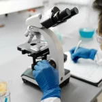 Peneliti bersarung tangan biru mengamati sampel melalui mikroskop di meja laboratorium