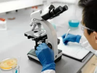 Peneliti bersarung tangan biru mengamati sampel melalui mikroskop di meja laboratorium