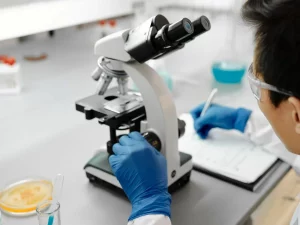 Peneliti bersarung tangan biru mengamati sampel melalui mikroskop di meja laboratorium